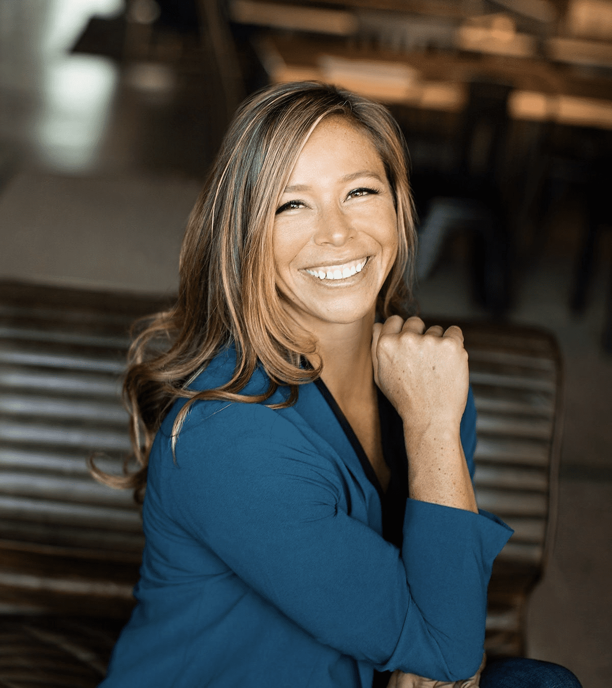 HeadshotPhoto Smiling person with long hair wearing a blue jacket, seated indoors with wooden furniture in the background.